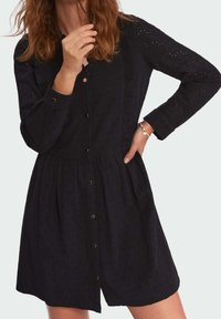 Mujer con un vestido negro de manga larga y botones, con un patrón texturizado, una mano en la cadera y la otra cerca de su rostro, llevando una pulsera.