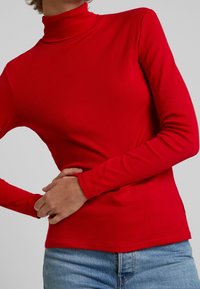 Czerwona, długorękawowa koszulka z golfem wykonana z miękkiej tkaniny, o dopasowanym kroju i przeszyciach dekoracyjnych. Zestawiona z jasnoniebieskimi dżinsami.