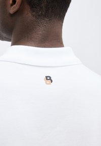 Vue de dos d'une personne portant un polo blanc avec un petit logo abstrait noir et beige sous le col.