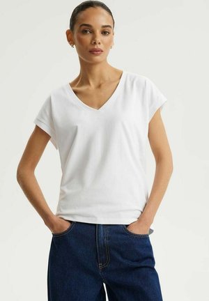 Kobieta z włosami związanymi do tyłu, ubrana w biały krótki rękaw z dekoltem w serek i ciemnoniebieskie jeansy, stojąca z rękami w kieszeniach.