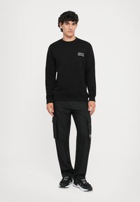 Sweatshirt preto com decote redondo e pequeno logo branco, combinado com calças cargo pretas com vários bolsos. Usado com ténis pretos.
