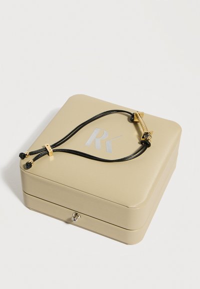 Μπεζ κουτί κοσμημάτων με ασημί λογότυπο "RK", που περιλαμβάνει ένα βραχιόλι με μαύρο κορδόνι και κούμπωμα σε σχήμα βίδας από χρυσό, το οποίο ξεκουράζεται στην κορυφή.