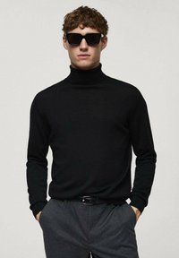 Schwarzer Rollkragenpullover aus Strickstoff, taillierte Passform, gerippte Bündchen. Getragen mit grauen Hosen und schwarzen Sonnenbrillen.