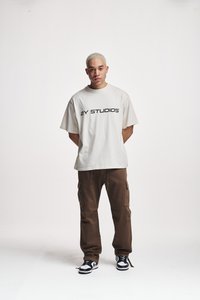 T-shirt oversize beige con testo "2Y STUDIOS" in nero, abbinata a pantaloni cargo marroni e sneaker bianco e neri. Sfondo neutro.