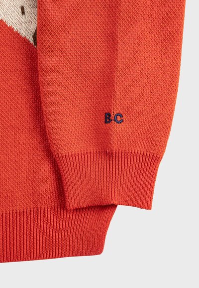 Gros plan sur la manche d'un pull orange en tricot avec un poignet côtelé et un petit détail de broderie "B.C" en bleu marine.