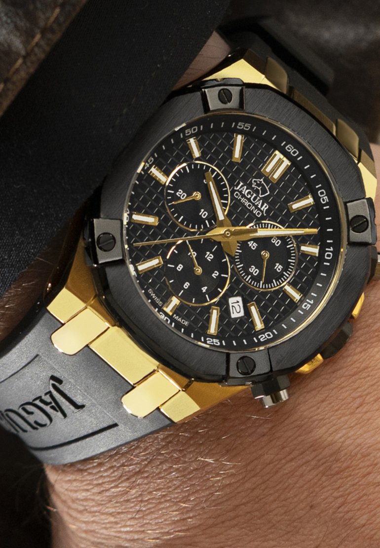 Reloj cronógrafo negro y dorado con una esfera negra texturizada, detalles plateados y una correa de goma gris con marca grabada.