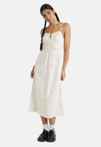 Vestido blanco de flores con tirantes finos, cuerpo fruncido y largo hasta la mitad de la pantorrilla. Presenta pequeñas flores de colores y un corte holgado, combinado con zapatos negros.
