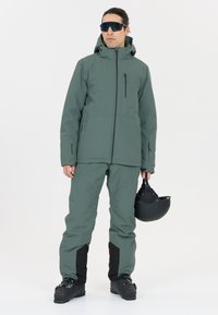 Skidjacka och byxor i dämpad grön färg med texturerad yta, med avtagbar huva, sidofickor och svarta detaljer. En svart skidhjälm hålls.
