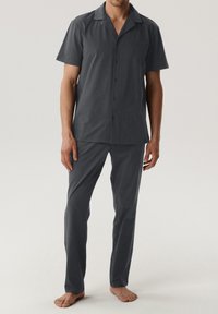 Ciemnoszara koszulka z krótkim rękawem, z zapięciem na guziki i kołnierzykiem, w połączeniu z dopasowanymi, szytymi na miarę spodniami; materiał wydaje się gładki i lekki.