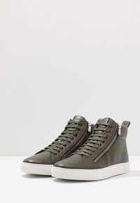 Hoge sneakers in donkergroen leer met rits- en vetersdetails, witte rubberen zool, gestructureerde afwerking en logo-accenten.
