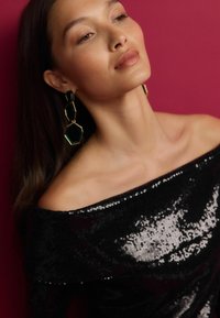 Sort off-shoulder bluse med perler, der har en glat tekstur. Store grønne geometriske øreringe med guld accents, sat mod en pink baggrund.