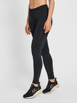 Legging noir slim avec poche latérale porté par une personne debout, associé à des baskets de sport noires et blanches.