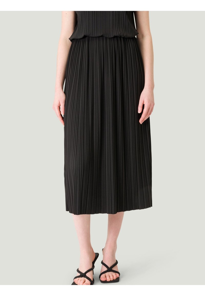 Femme portant une jupe midi plissée noire et des sandales noires à lanières, debout devant un fond gris clair uni.