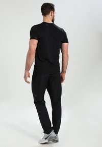 Hombre con barba, vistiendo camiseta negra, pantalones negros y zapatillas Nike blancas, de pie y de espaldas contra un fondo liso.