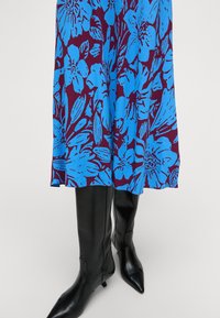 Gėlių suknelė su ryškiu mėlynu raštu tamsiai purpuriniame fone, derinama su elegantiškais juodais aštriu nosiu batais.