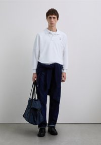 Polo blanc à manches longues, pull en tricot bleu marine noué à la taille, pantalon en jean foncé, chaussures noires, tenant un sac sombre texturé.