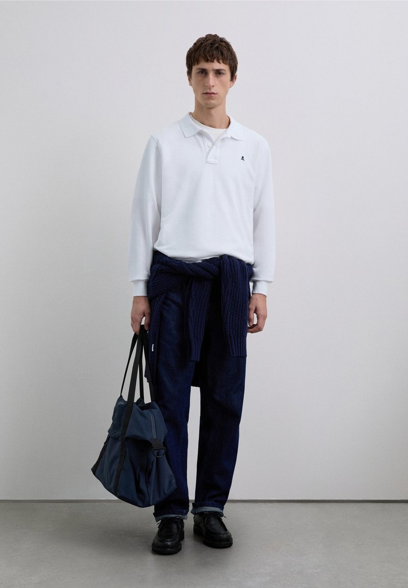 Polo blanc à manches longues, pull en tricot bleu marine noué à la taille, pantalon en jean foncé, chaussures noires, tenant un sac sombre texturé.