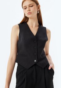 Schwarze Weste mit V-Ausschnitt, tailliertem Design, Fünf-Knopf-Verschluss und Seitentaschen, aus glattem Stoff gefertigt. Minimalistischer Stil.