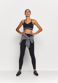 Black sports bra with white logo, high-waisted black leggings, gray sweatshirt tied around the waist, and black athletic shoes with white accents.