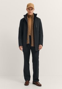 Schwarzer wasserdichter Mantel, brauner gemusterter Rollkragenpullover, dunkle Hose und braune Mütze, mit braunen Lederschuhen; verfügt über eine mit Reißverschluss versehenen Brusttasche.