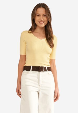 Smilende kvinde med langt brunt hår iført en gul kortærmet ribstrikket top, hvide højtaljede bukser og et mørkebrun bælte.