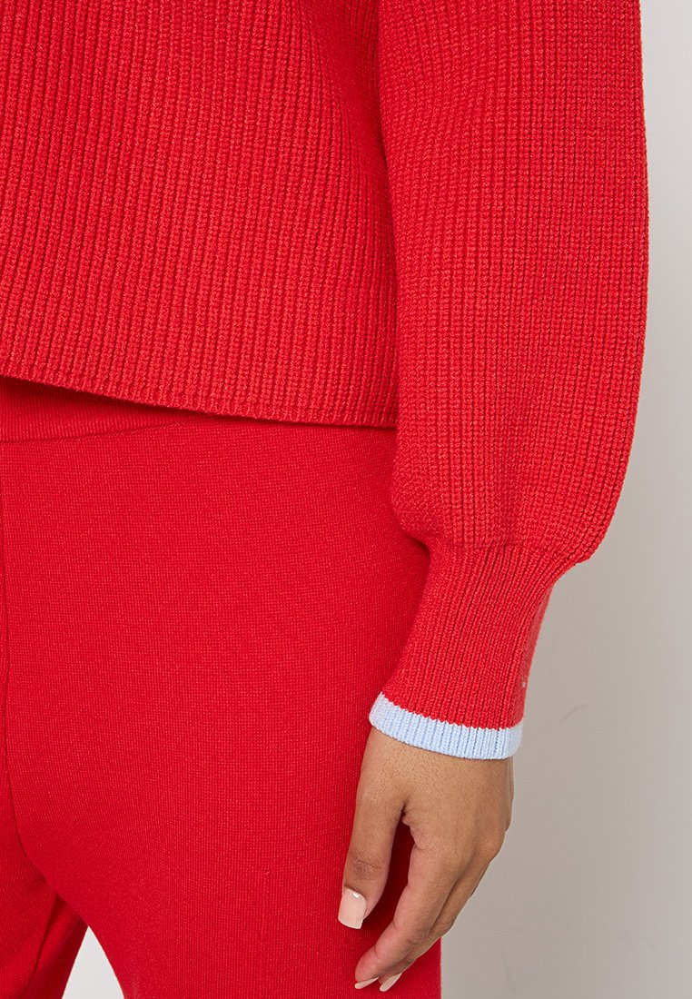 Nahaufnahme einer Person, die einen roten Strickpullover und passende Hosen trägt, die Hand entspannt an ihrer Seite, mit einem hellblauen Bunddetail.