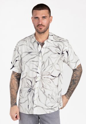 Mężczyzna o krótkich włosach i tatuażach na obu ramionach nosi białą, krótkorękawową koszulę zapinaną na guziki z dużymi czarnymi i szarymi kwiatowymi wzorami.