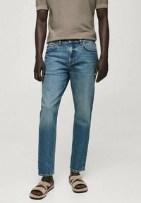 Mann trägt mittelblaue gerade Jeans, ein beiges kurzärmliges Strickoberteil und beigen Sandalen vor einem schlichten hellen Hintergrund.