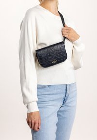Sac à bandoulière noir en cuir à effet croco avec un rabat courbé et un accent logo doré. Porté sur une sangle d'épaule, associé à un pull blanc et un jean bleu.