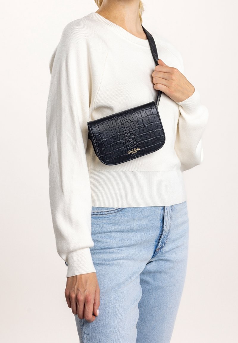 Sac à bandoulière noir en cuir à effet croco avec un rabat courbé et un accent logo doré. Porté sur une sangle d'épaule, associé à un pull blanc et un jean bleu.