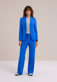 Jasnofialový oblek so šitým sako a nohavicami s širokými nohavicami, doplnený šedým topom; hladká látka a štruktúrovaný dizajn.