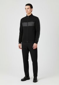 Czarna kurtka z suwakiem, z teksturowanym czarnym panelem na wysokości klatki piersiowej, zestawiona z czarnymi, dopasowanymi spodniami oraz czarnymi mokasynami. Gładka tkanina, minimalistyczny design.