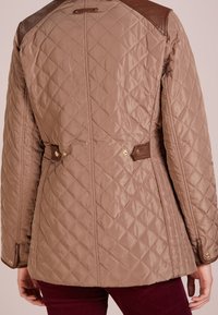 Prošívaná béžová bunda s hnědými koženými ramenními akcenty, bočními přezkami a zlatými detaily na manžetách. Hladká textura a přiléhavý střih.
