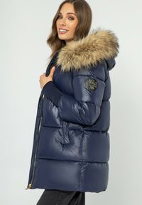 Marineblauwe gewatteerde jas met een nepbont capuchon, ritssluiting en logo patch op de mouw. Kenmerkt zich door een gewatteerd ontwerp en zijzakken.