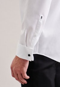Camicia bianca in tessuto liscio, con un bottone decorativo nero e un dettaglio ricamato di rosa nero sul polsino.
