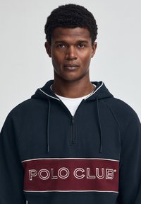 Sudadera con capucha azul marino con media cremallera, cordón blanco y una franja granate con "POLO CLUB" bordado en blanco en el frente.