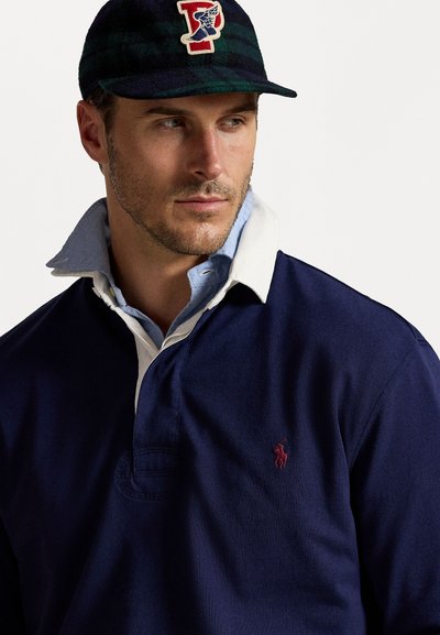 Cămașă polo din bumbac navy cu guler alb și logo brodat. Asortată cu o șapcă cu carouri negre și verzi închise, având un patch cu logo.