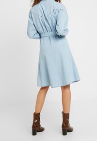 Light blue denim shirt dress with a belted waist, long sleeves, and decorative studs on the back yoke. Brown glossy heeled boots.