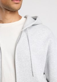 Sudadera con cremallera de color gris claro con una textura suave, que presenta una capucha con cordón y un logo sutil en el pecho. Lleva puesta sobre una camiseta de color crema.