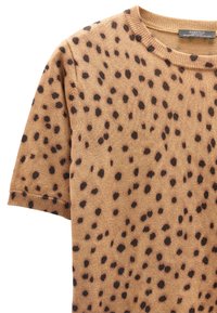 Brązowy sweter z krótkim rękawem, pokryty nieregularnymi czarnymi plamkami, z okrągłym dekoltem, ukazujący górną część przodu i rękaw.
