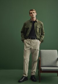 Chaqueta de tela verde sobre una camiseta negra, combinada con pantalones cargo beige y zapatos negros de cordones. Silla gris en el fondo.