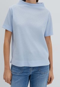 Top a maniche corte in azzurro chiaro, realizzato in tessuto testurizzato, con colletto alto e vestibilità rilassata, abbinato a jeans azzurro chiaro.