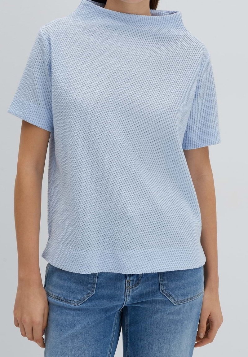 Top a maniche corte in azzurro chiaro, realizzato in tessuto testurizzato, con colletto alto e vestibilità rilassata, abbinato a jeans azzurro chiaro.