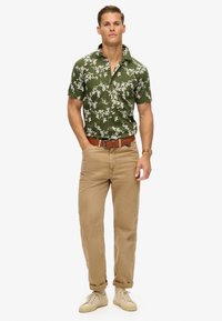 Korte mouwen, groene bloemenhemel met een knoopkraag, gedragen met beige broeken en neutrale sneakers, die een ontspannen pasvorm en textuur laat zien.
