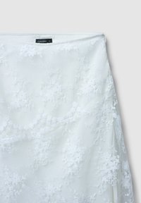 Weißer Spitzenrock mit floraler Stickerei, transparenter Textur und glatter Stoffoberfläche. Enthält ein schwarzes Etikett mit Markennamen.