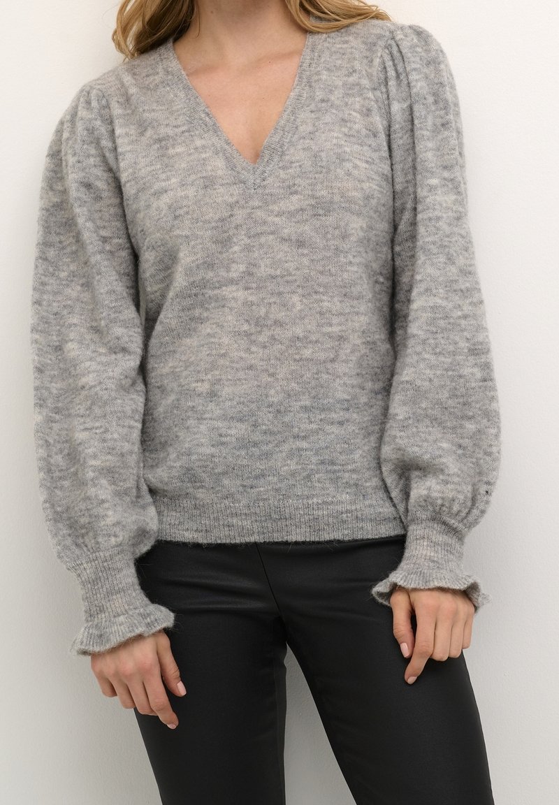 Szary, miękki sweter z dekoltem w kształcie V, bufkami oraz falbaniastymi mankietami, wykonany z teksturowanej dzianiny. Zestawiony z czarnymi spodniami.