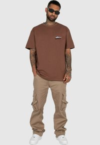 T-shirt en coton marron avec un petit graphique bleu et noir sur la poitrine gauche, associé à un pantalon cargo beige et des baskets blanches.