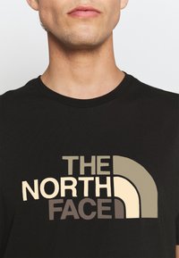 Black t-shirt featuring a multicolored graphic logo with "THE NORTH FACE" in beige and earthy tones. Crew neck design, short sleeves.