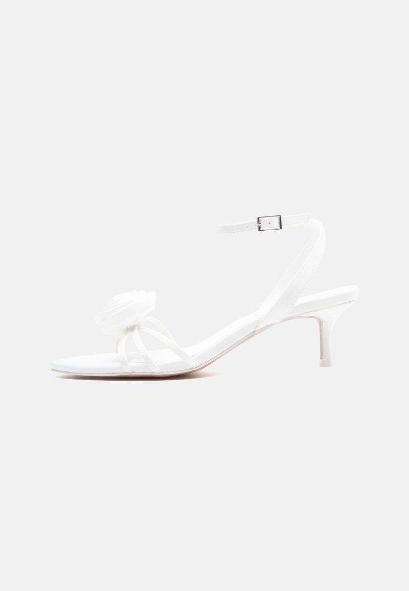 Λευκό σανδάλι με λουράκια, χαμηλό τακούνι kitten, λουράκι με πόρπη στον αστράγαλο και διακοσμητικό λουλούδι στο μπροστινό λουράκι.