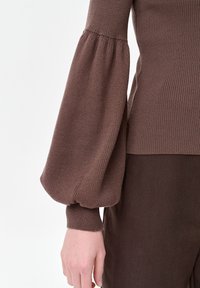 Bruine geribde gebreide trui met volumineuze ballonmouw die bij de pols is samengebonden, gecombineerd met een donkerbruine broek aan een lichtgekleurde arm.